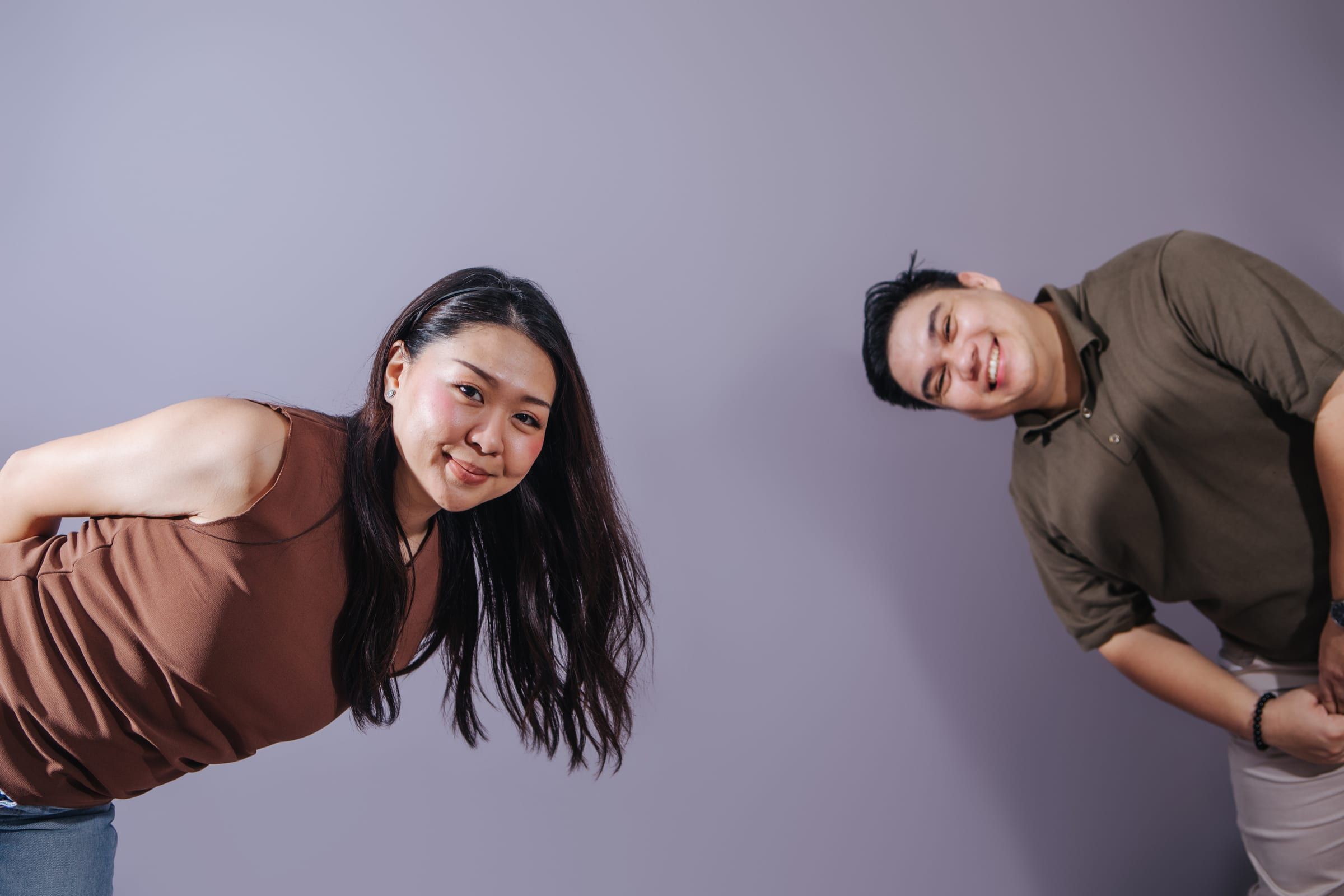 Anna and Joboy lean in from opposite edges of the frame, grinning at the camera across an empty stretch of lavender wall.