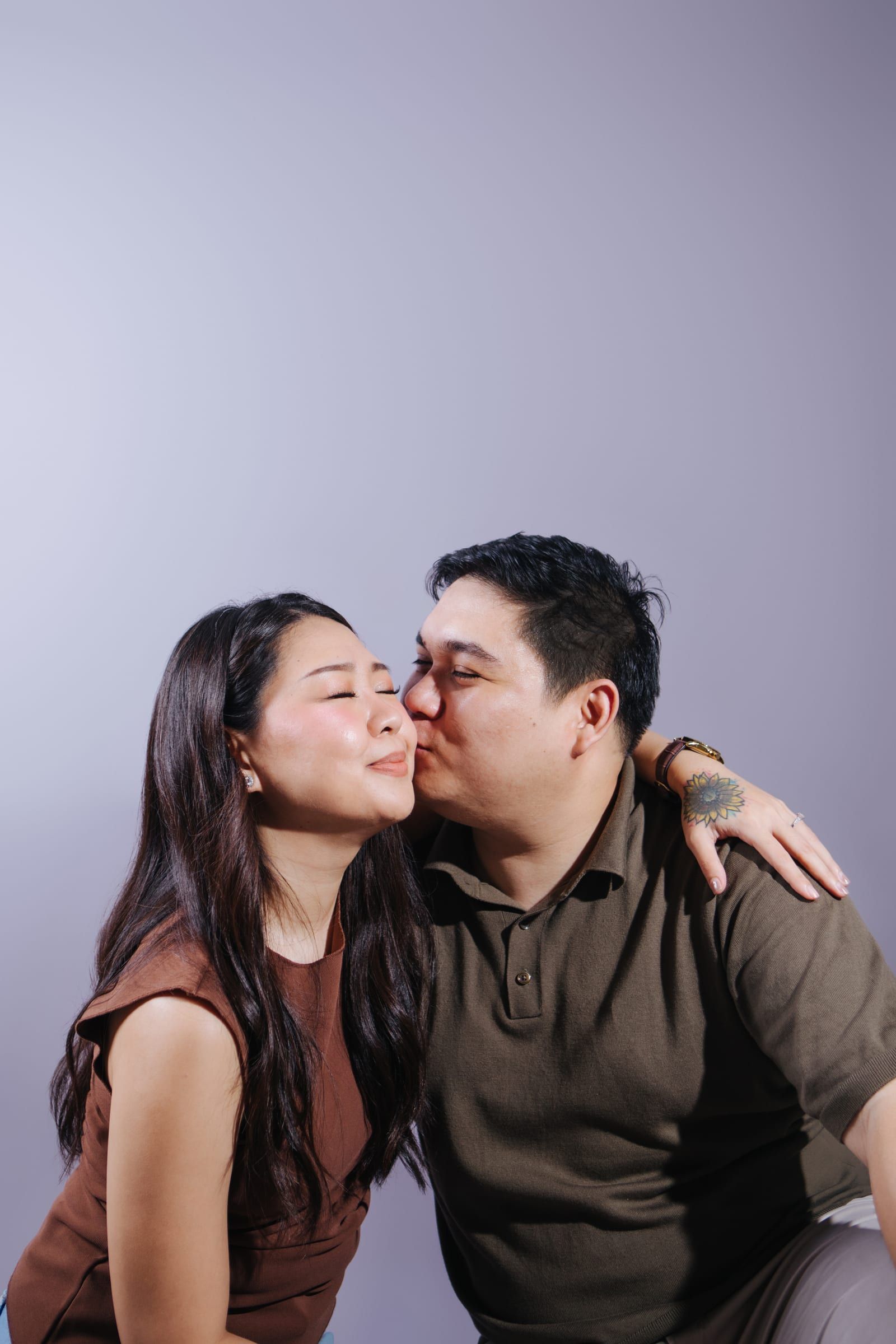 Joboy kisses Anna on the cheek while she closes her eyes and smiles, his arm around her shoulders, with open lavender backdrop above them.
