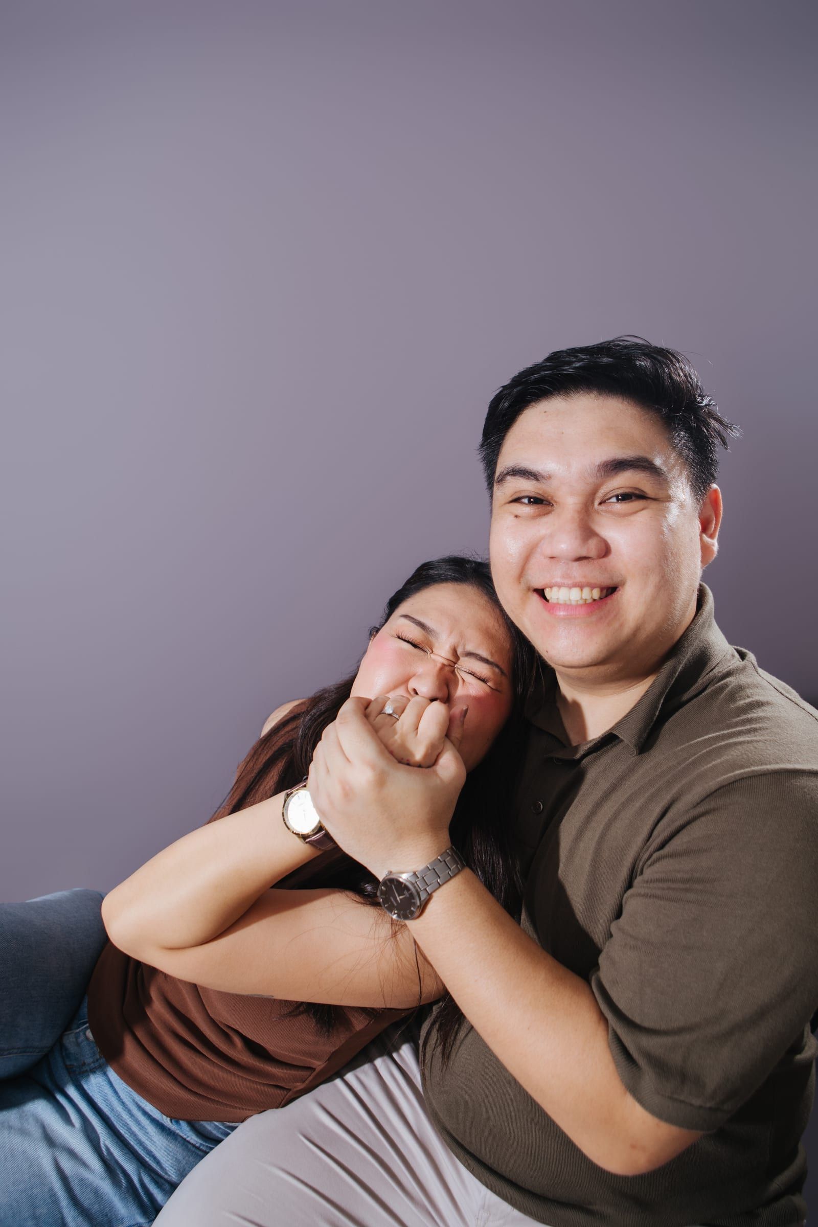 Anna laughs with her eyes squeezed shut behind their joined hands, her engagement ring showing, while Joboy grins at the camera against a lavender backdrop.